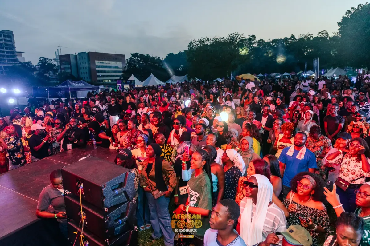 A crowd of several hundred people at dusk facing the main stage, with white festival marquees and food stalls behind them