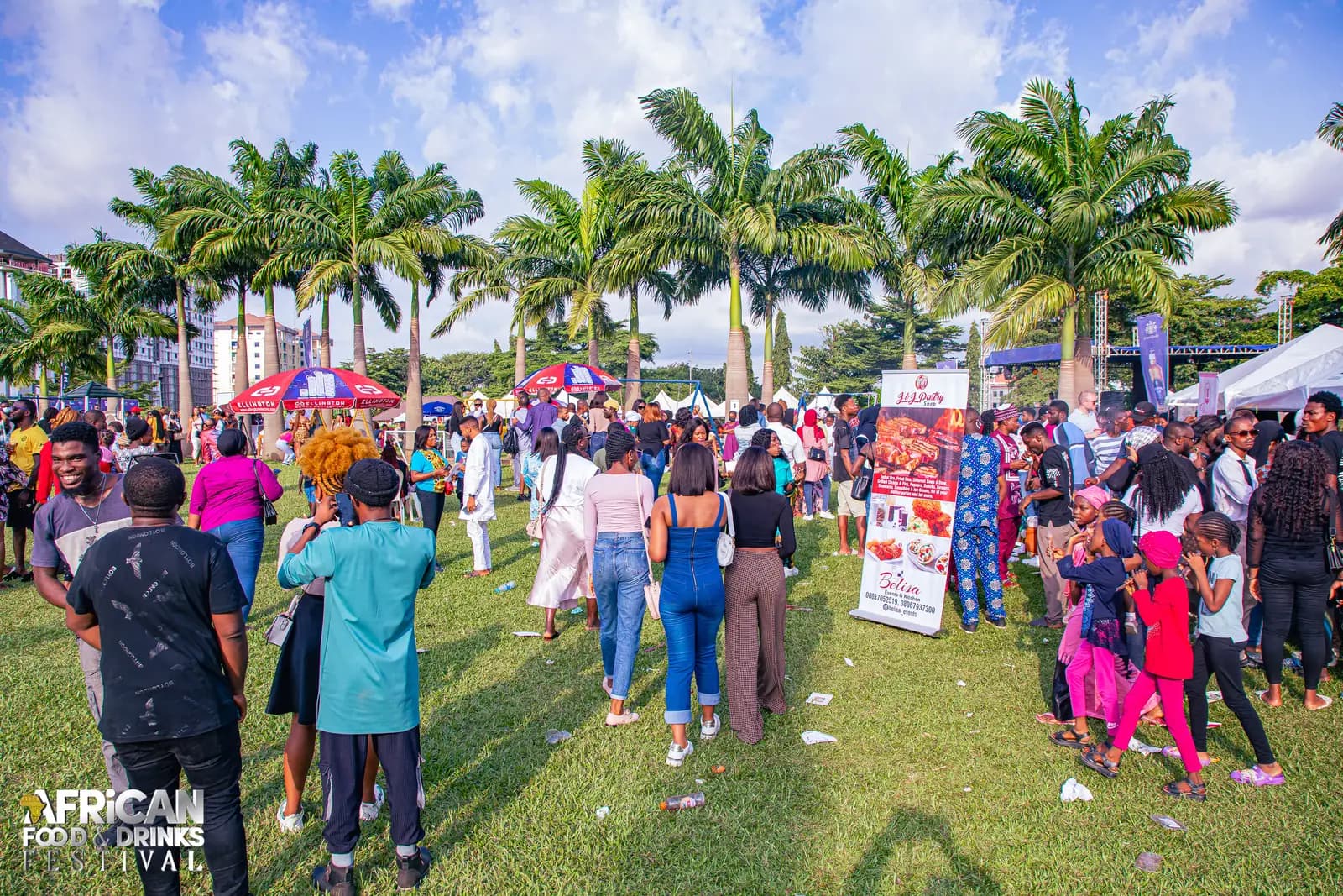 Une foule en plein jour sur une pelouse bordée de grands palmiers, les bannières et les stands du festival au loin sous un ciel bleu