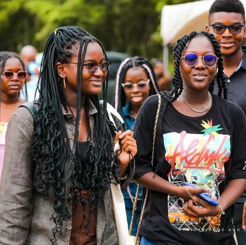 Trois invitées ensemble en plein air, l'une en tee-shirt imprimé Accra, la foule derrière elles