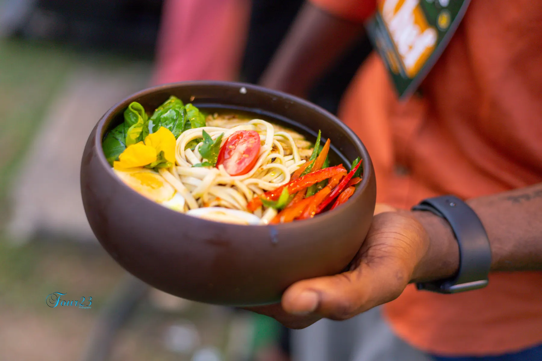 Un bol en bois de soupe de nouilles avec tomate, piment et une fleur jaune, tendu vers l'objectif