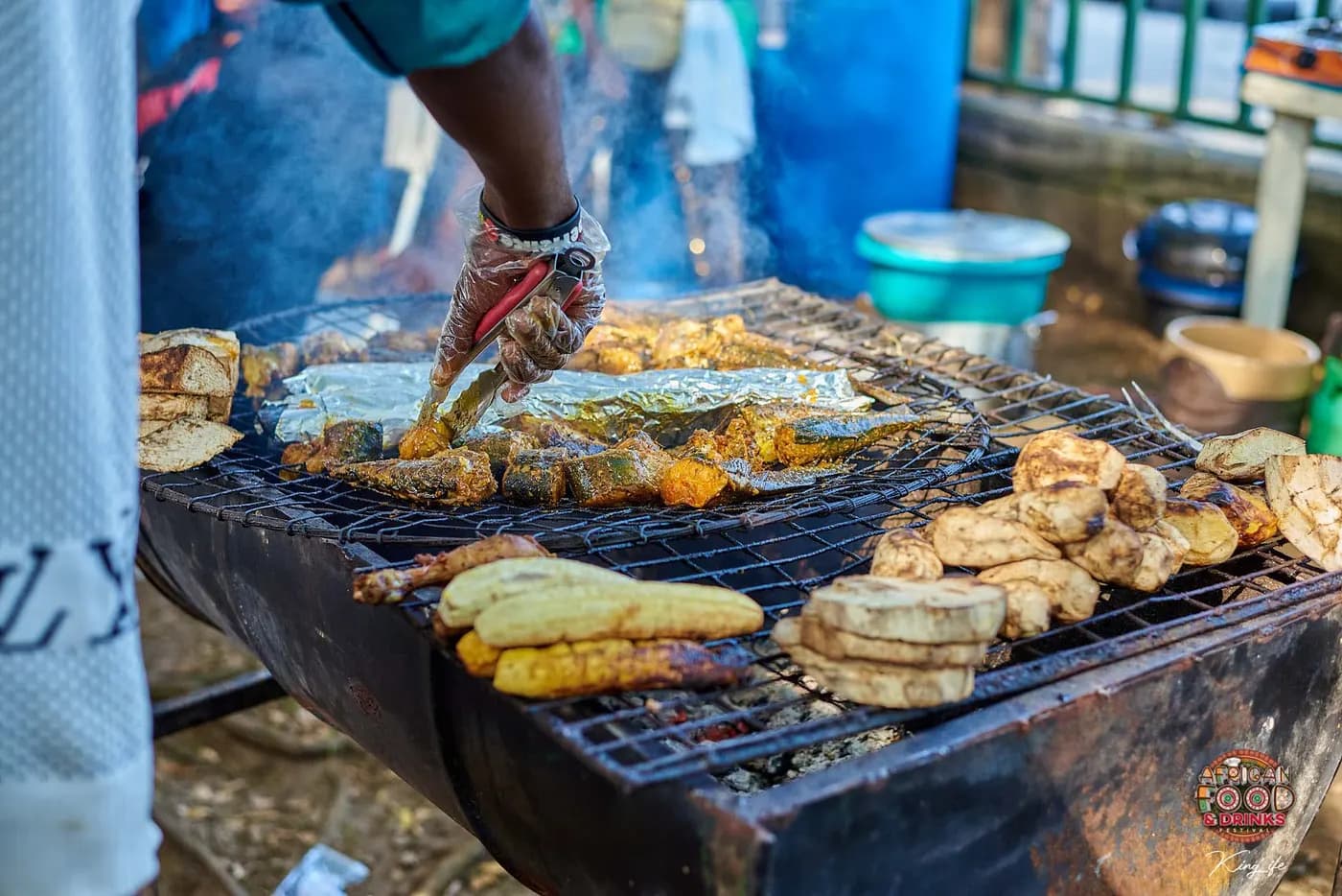 Une main gantée retourne du maquereau épicé avec des pinces sur un gril à charbon, plantain et igname cuisant à côté