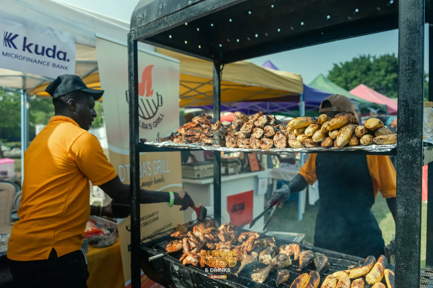 Deux vendeurs en chemise orange s'affairent sur un double gril chargé de poulet braisé et de plantain grillé, les chapiteaux du festival derrière