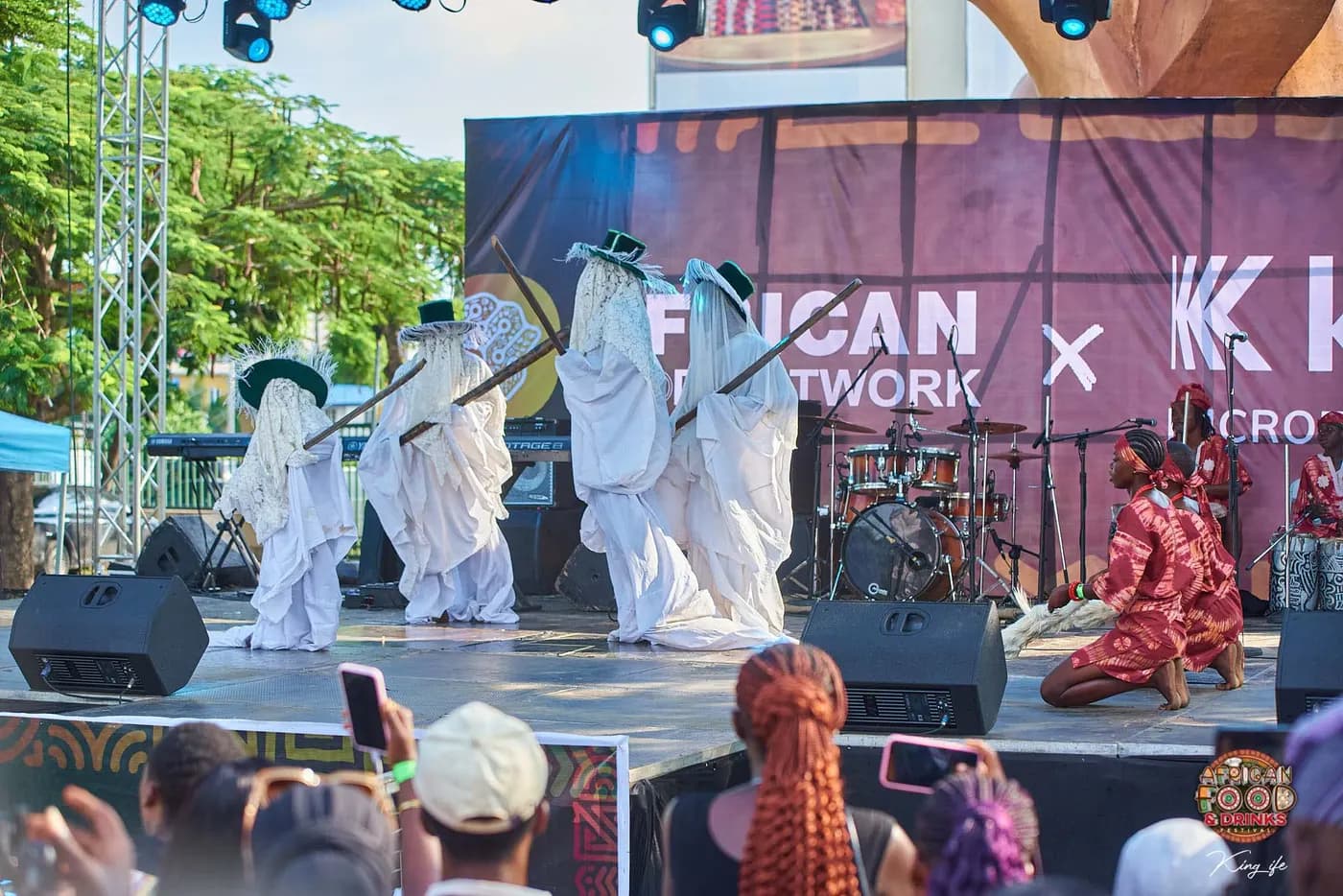 Quatre danseurs masqués en robes blanches et chapeaux à franges sur scène devant une bannière African Food Network, les percussionnistes sur le côté