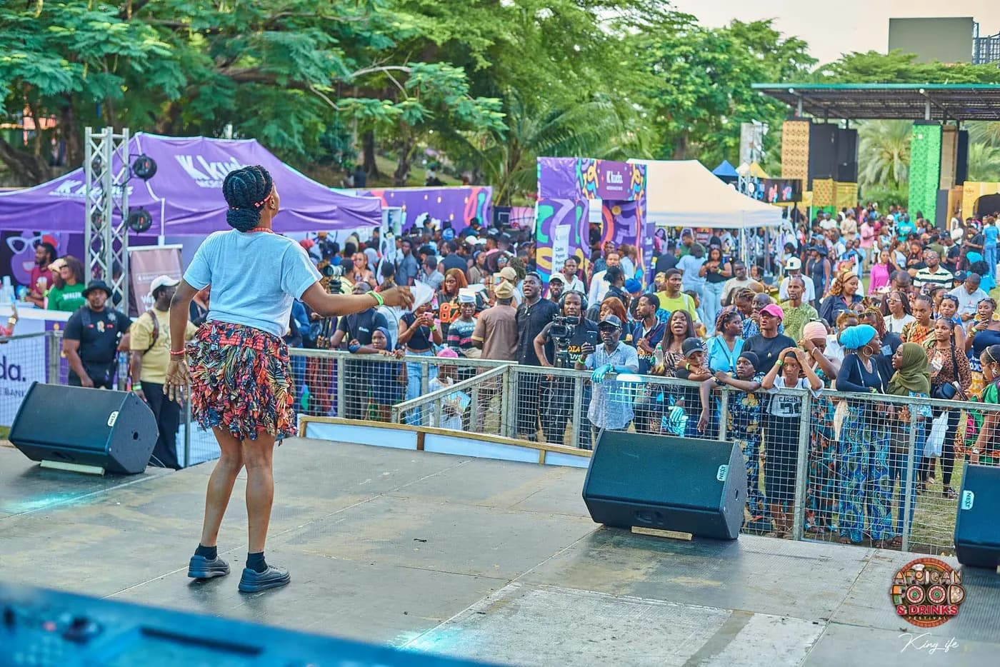 Une artiste au bord de la scène tend la main vers un public massé contre la barrière, les stands du festival derrière